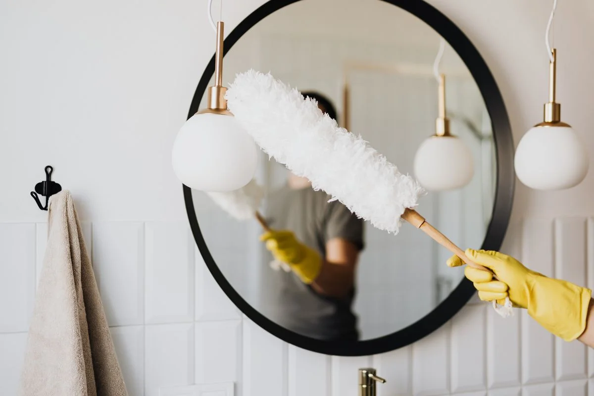 A gloved cleaner dusting a bathroom mirror with a white duster